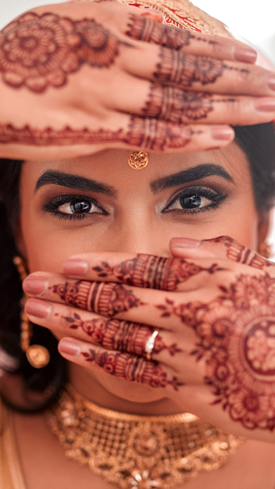 Bride showcasing intricate mehndi designs on her hands with captivating eye makeup and traditional gold jewelry, capturing a close-up wedding moment.